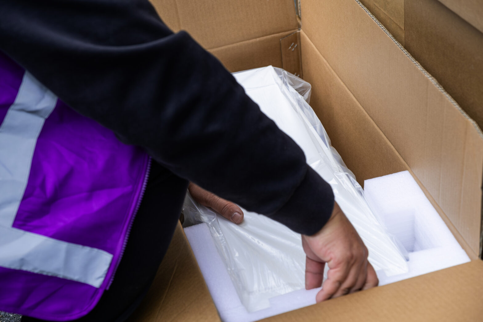 A person in a purple vest is unpacking a white item from a cardboard box, surrounded by protective packaging.