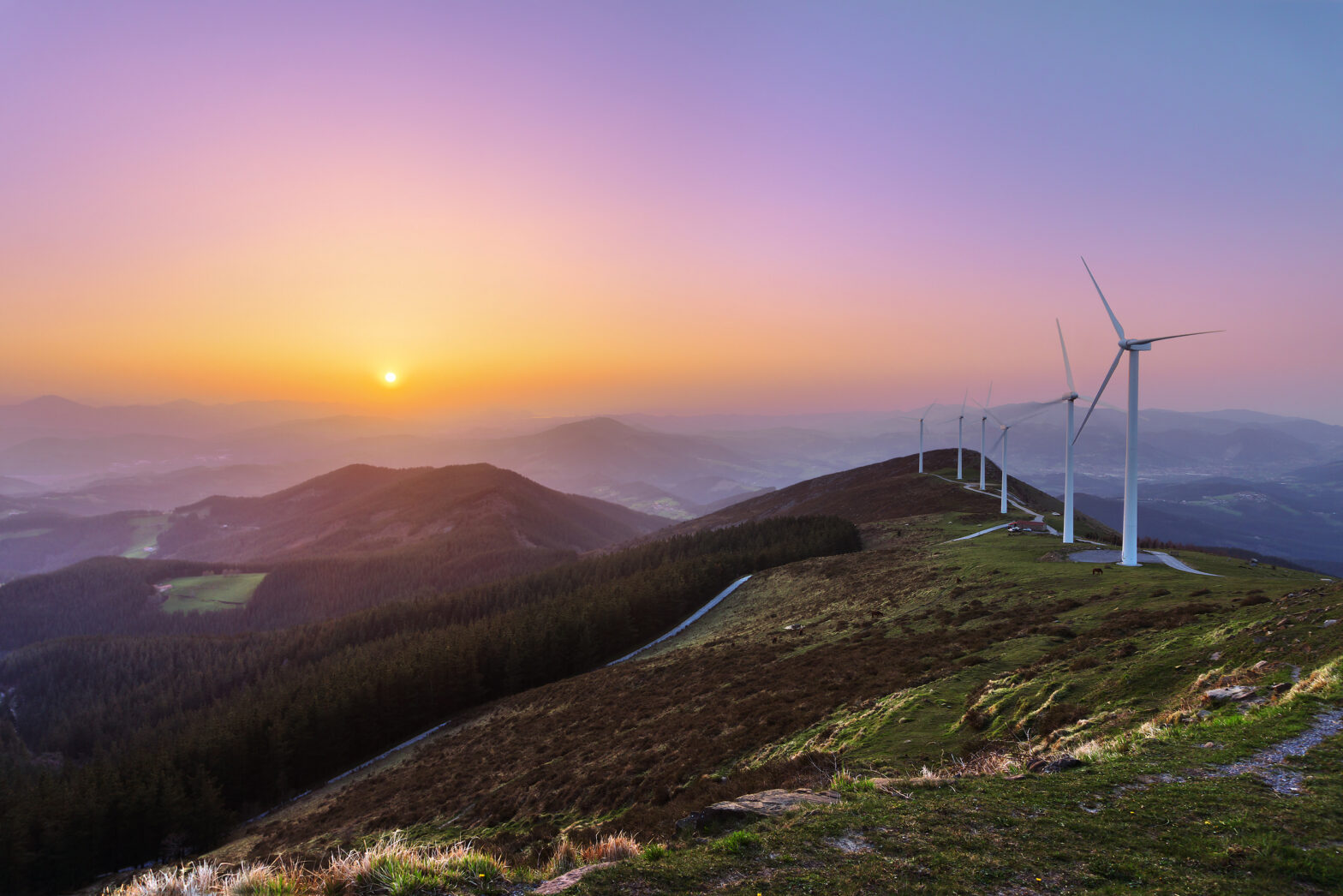 A scenic view of wind turbines on a hillside at sunset, with rolling mountains and a colorful sky in the background.