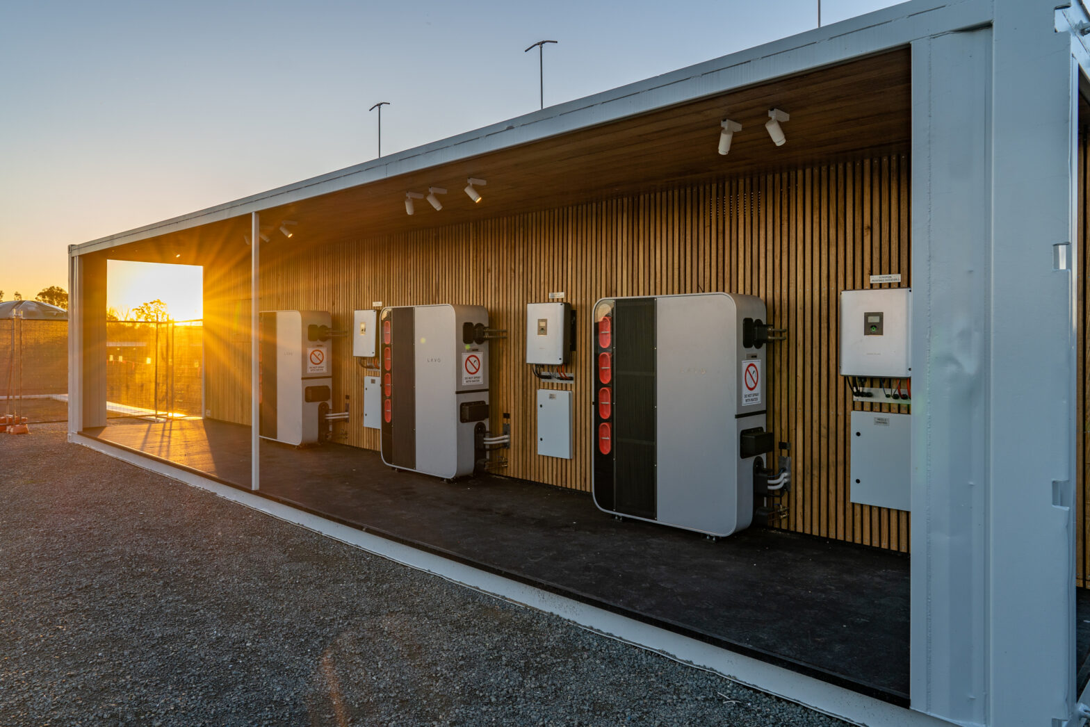 A modern charging station with multiple units illuminated by the sunset, set against a wooden backdrop.